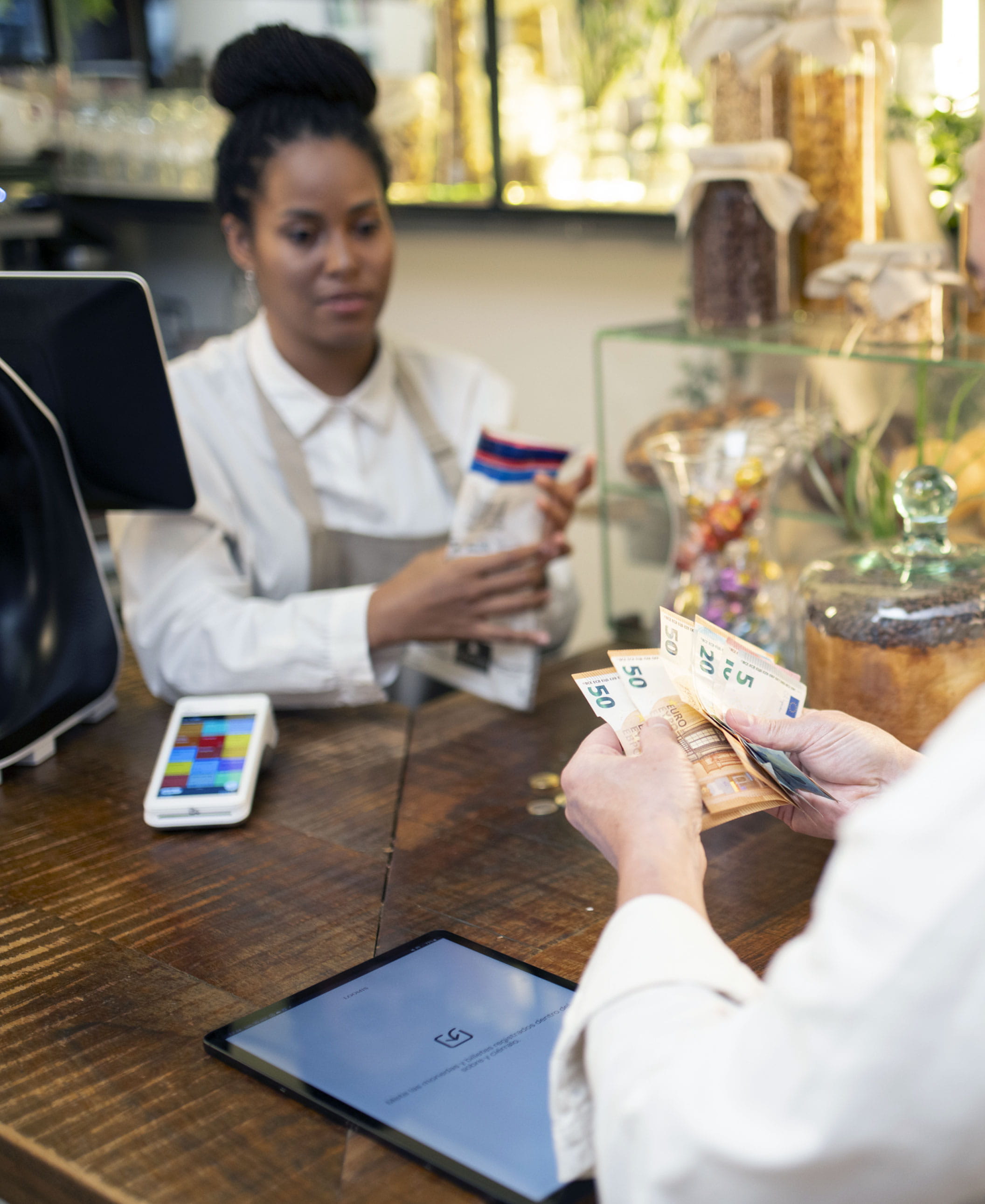 A la derecha, dos manos con billetes sobre una barra. Al otro lado, una chica con un sobre en la mano. Frente a ella, el dispositivo Pay-Mobile Loomis Pay con el programa de gestión y una parte del dispositivo Pay-Desk al lado.
