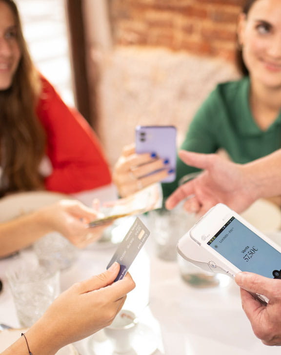 Distintas manos sobre una mesa: a la izquierda el Pay-Mobile Loomis Pay, seguido de un iPhone, efectivo y una tarjeta de crédito.