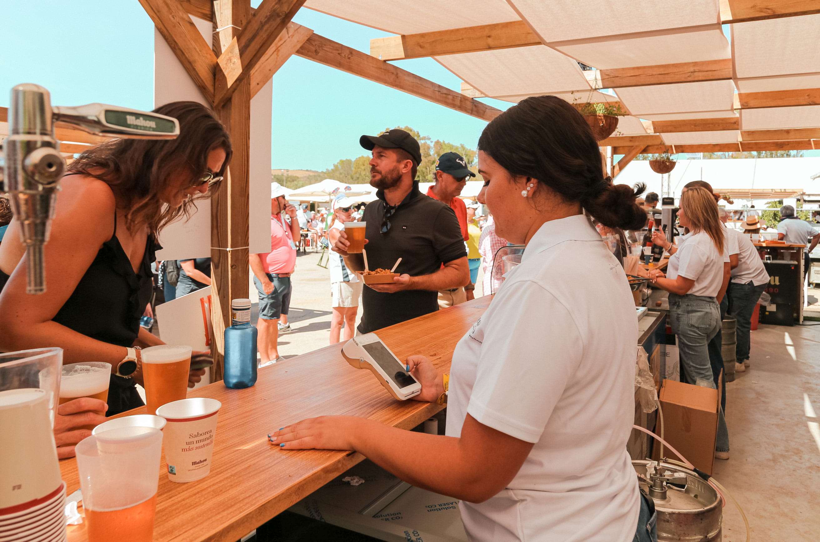 Chica detrás de la barra mirando el Pay-Mobile Loomis Pay, mientras otra chica simula buscar algo para pagar.