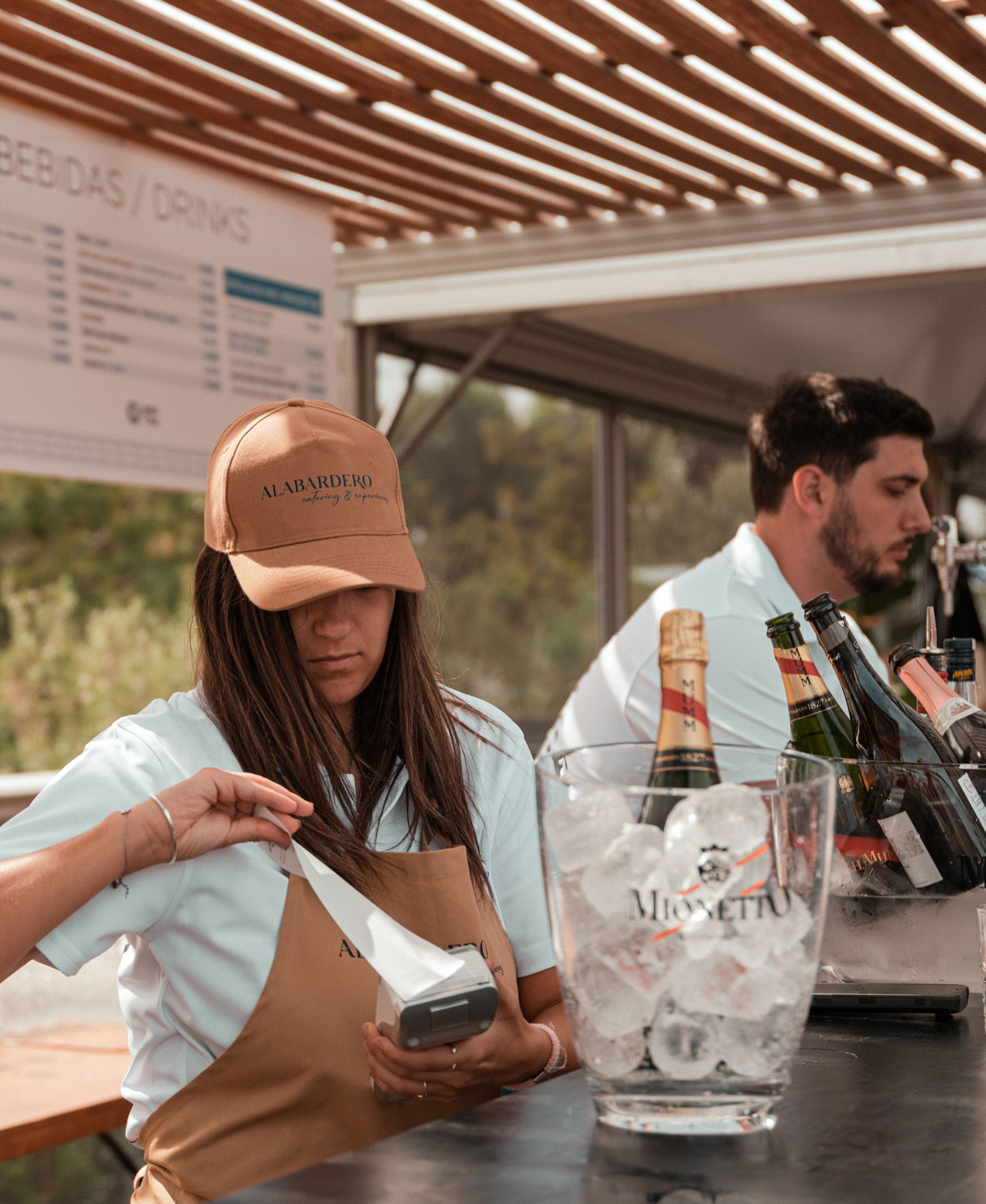Dos camareros detrás de la barra, una chica con gorra sacando un ticket del Pay-Mobile Loomis Pay. En la mesa, una cubitera con champán y otra detrás con más botellas.
