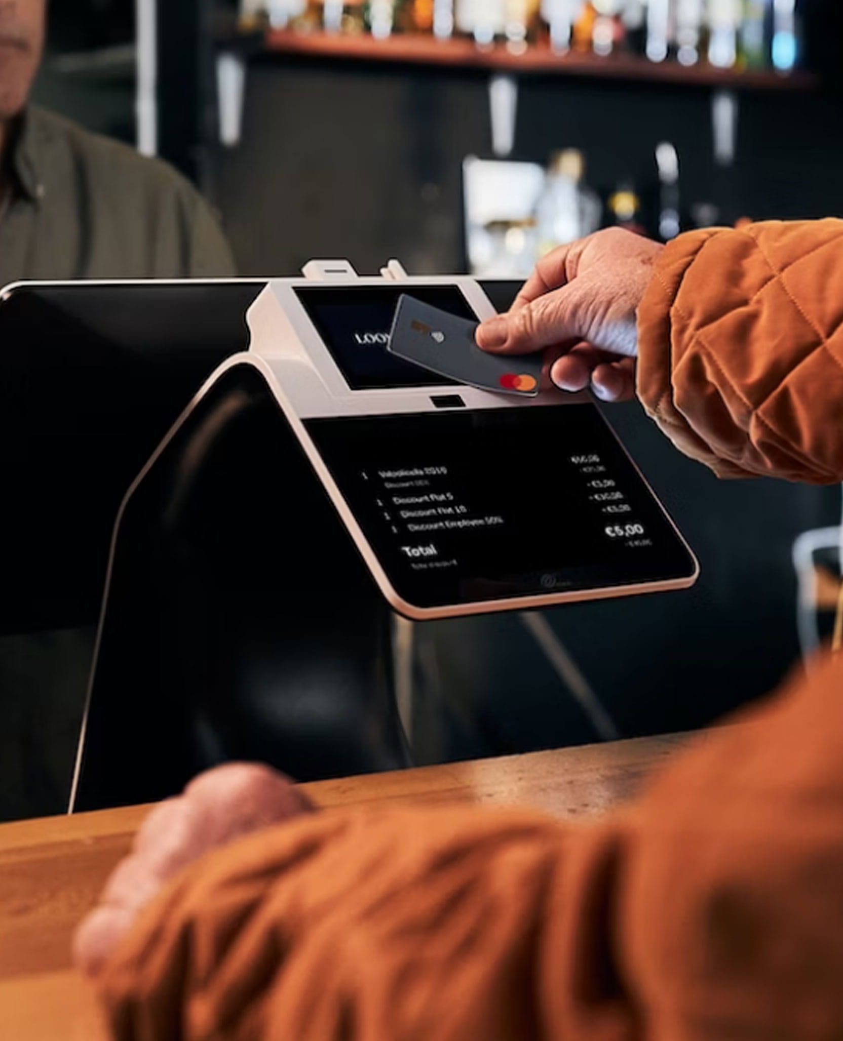 Fotografía del dispositivo Pay-Desk Loomis Pay detrás de una mesa alta, con un brazo a la derecha acercando una tarjeta para generar el pago. En la pantalla se muestra el pedido a pagar, con el nombre de los productos y su precio.