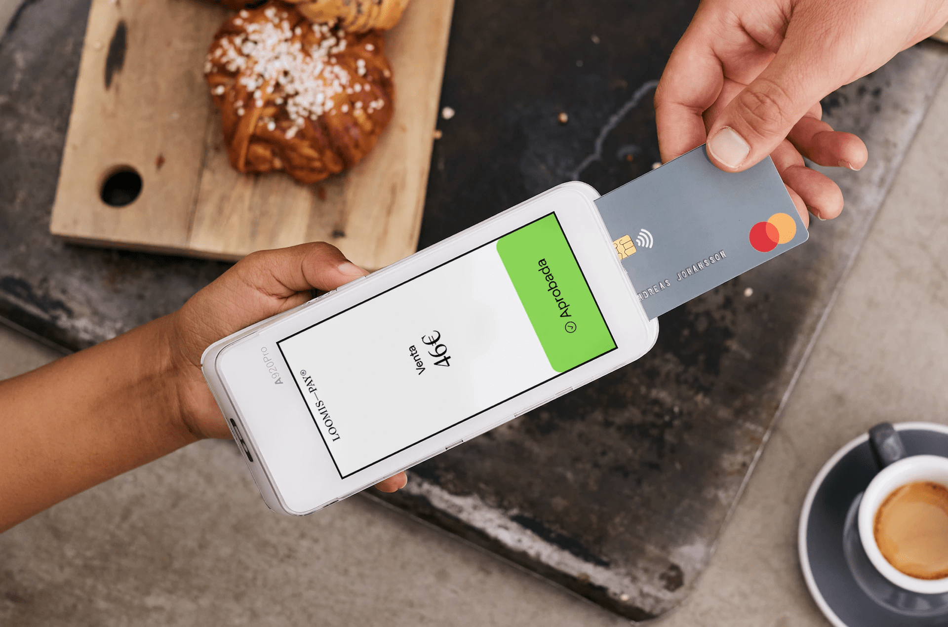 Primer plano del Pay-Mobile Loomis Pay con tarjeta de crédito insertándose y pantalla mostrando la aprobación de un pago de 46 euros. Al fondo, una mesa negra y otra de madera con dos bollos.