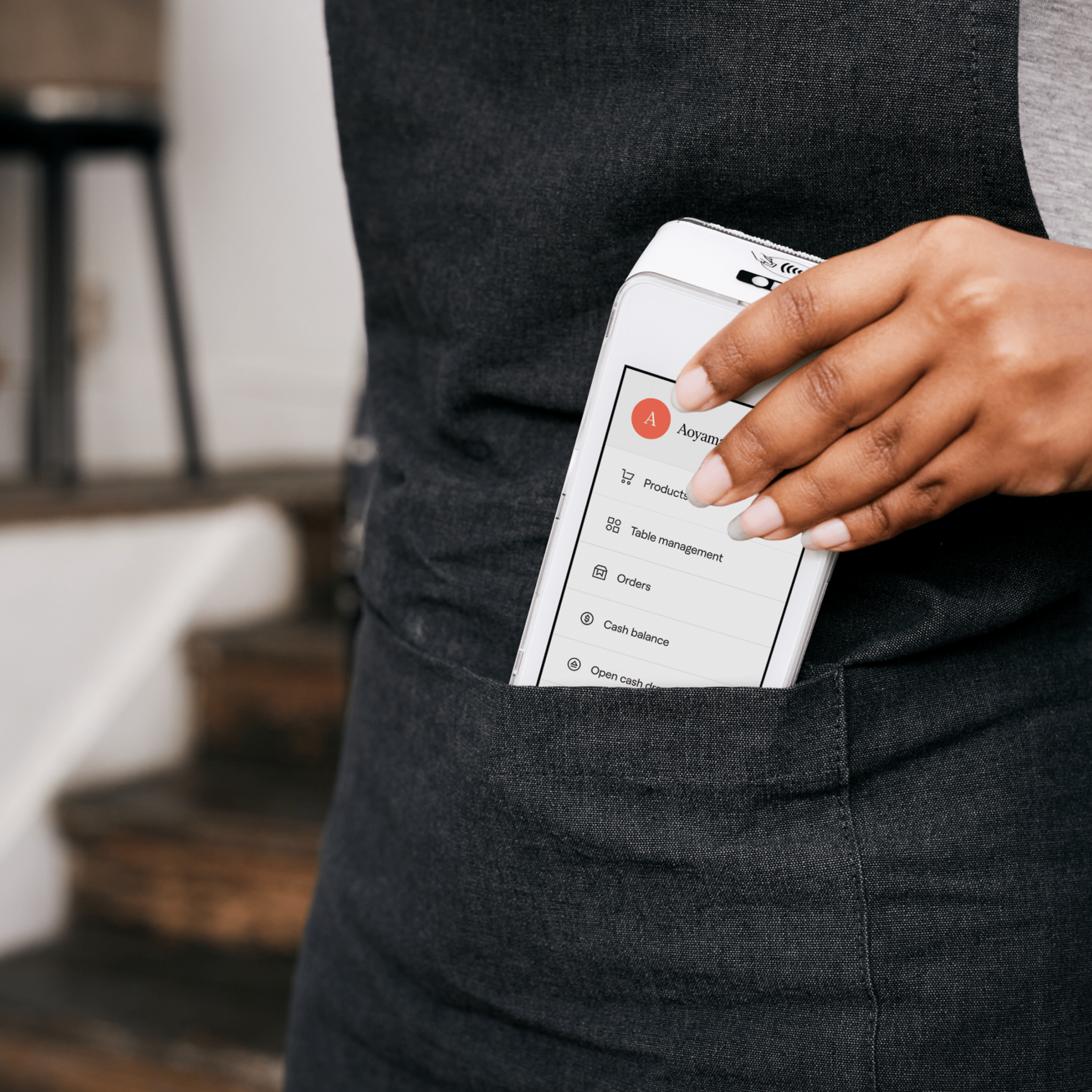 Primer plano de un dispositivo A920 con el software Loomis Pay, sostenido desde arriba por la mano de una persona, simulando que lo extrae del bolsillo de su delantal.